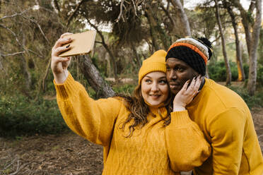Junge Frau mit Strickmütze, die ein Selfie mit ihrem Mobiltelefon macht, während sie im Wald steht - EGAF01384