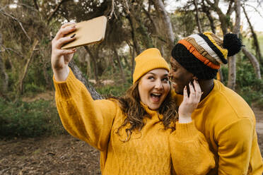 Fröhliche Frau, die ein Selfie mit ihrem Mobiltelefon macht, während sie im Wald steht - EGAF01386