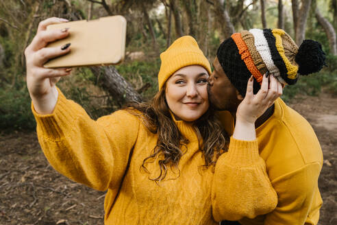 Mann küsst Frau, die ein Selfie mit ihrem Handy macht, während sie im Wald steht - EGAF01387