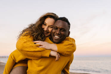 Smiling man piggybacking woman while standing at beach - EGAF01396