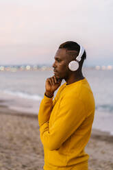 Mittlerer erwachsener Mann mit Kopfhörern, der Musik hört, während er am Strand steht - EGAF01405
