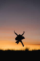 Carefree man wearing jacket jumping with arms outstretched against sky during sunset - EGAF01411