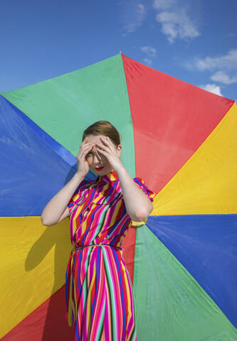 Woman shielding eyes while standing against colorful beach umbrella stock photo