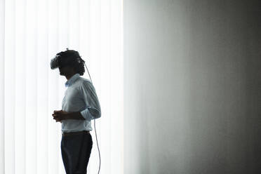 Junger Unternehmer mit Virtual-Reality-Headset vor einem weißen Vorhang im Büro - JOSEF02840