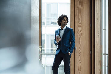 Thoughtful young handsome businessman holding smart phone while looking away against window at office - JOSEF02851