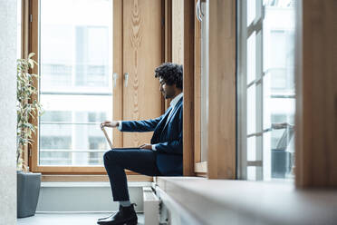 Young male entrepreneur using laptop while sitting against window at workplace - JOSEF02863