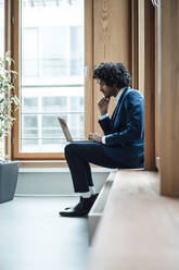 Young businessman using laptop while sitting against window at workplace - JOSEF02864