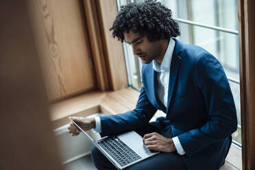Junge männliche Fachkraft, die einen Laptop benutzt, während sie im Büro am Fenster sitzt - JOSEF02865