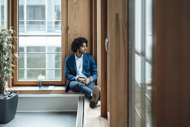 Thoughtful male entrepreneur looking away while sitting by laptop against window at office - JOSEF02866