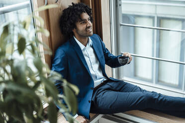 Lächelnder männlicher Fachmann, der wegschaut und eine Kaffeetasse hält, während er am Fenster im Büro sitzt - JOSEF02868