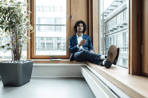 Confident male entrepreneur holding coffee cup while sitting against window at office - JOSEF02869