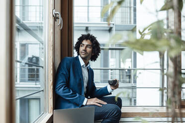 Thoughtful young male professional looking away while holding coffee cup sitting with laptop against window at office - JOSEF02871