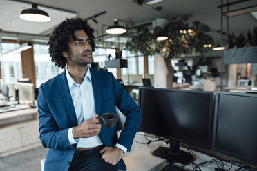 Nachdenklicher männlicher Unternehmer, der eine Kaffeetasse in der Hand hält, während er neben Computermonitoren am Schreibtisch im Büro sitzt - JOSEF02872
