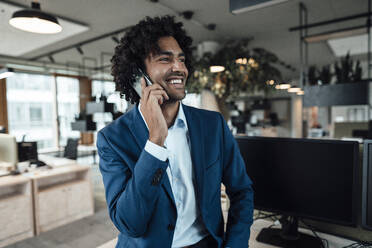 Lächelnder junger männlicher Unternehmer, der mit seinem Smartphone spricht und seinen Blick vom Arbeitsplatz abwendet - JOSEF02874