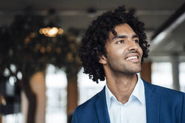 Smiling handsome young businessman with curly black hair looking away at office - JOSEF02888