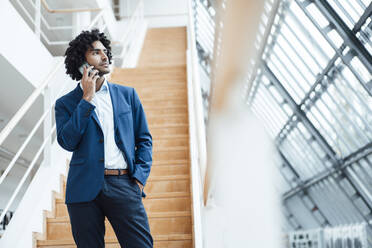 Handsome young male professional talking on mobile phone while standing against staircase in office - JOSEF02892