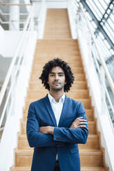 Confident handsome young male entrepreneur standing with arms crossed against staircase at office - JOSEF02893