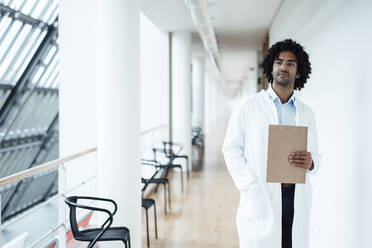 Nachdenklicher junger Mann im Gesundheitswesen, der ein Klemmbrett hält, während er in einem Krankenhaus auf dem Flur steht - JOSEF02901