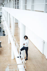 Smiling young businessman standing with arms crossed by column in office corridor - JOSEF02904
