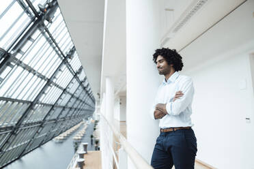 Young male professional with arms crossed leaning on white column in corridor at office - JOSEF02905