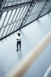 Young businessman with digital tablet walking by roof in office corridor - JOSEF02910