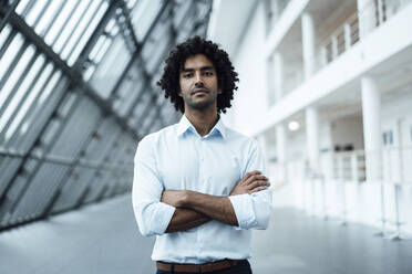 Confident young businessman standing with arms crossed at workplace - JOSEF02916