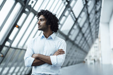 Thoughtful young male professional standing with arms crossed while looking away at workplace - JOSEF02917