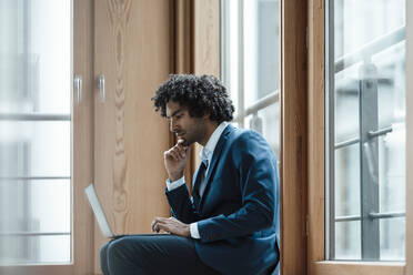 Junger männlicher Unternehmer, der einen Laptop benutzt, während er am Arbeitsplatz am Fenster sitzt - JOSEF02927