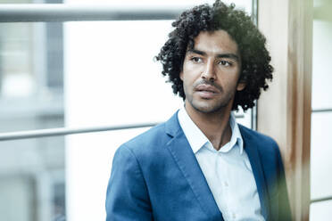 Thoughtful young businessman looking away while sitting against window at workplace - JOSEF02931