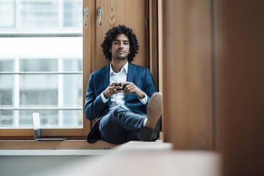 Confident male entrepreneur holding coffee cup while sitting against window at office - JOSEF02933