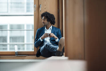 Young businessman holding coffee cup while looking away sitting against window at office - JOSEF02934