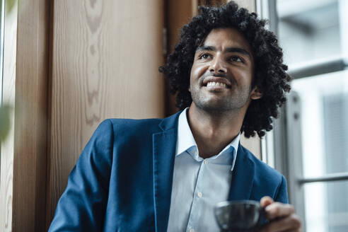 Lächelnder Geschäftsmann, der eine Kaffeetasse hält und gegen ein Fenster am Arbeitsplatz schaut - JOSEF02937