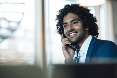 Smiling young male professional talking on landline phone while looking away at workplace - JOSEF02941