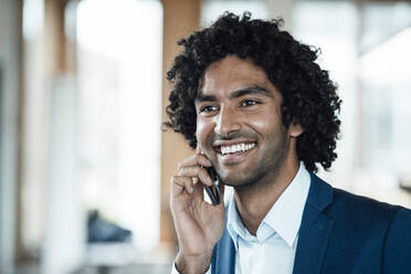Cheerful young businessman talking on mobile phone while looking away in office - JOSEF02943