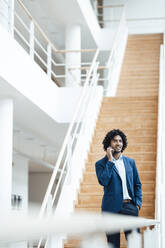 Young male professional talking on mobile phone while looking away against staircase in office - JOSEF02956