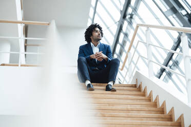 Thoughtful male entrepreneur looking away while sitting with hands clasped on staircase in office - JOSEF02963