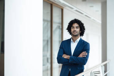 Handsome young male professional standing with arms crossed contemplating in corridor of office - JOSEF02965