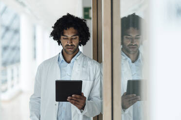 Young male doctor using digital tablet while leaning on glass wall at hospital - JOSEF02976