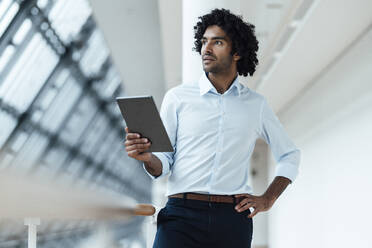 Nachdenklicher männlicher Unternehmer, der ein digitales Tablet in der Hand hält, während er mit der Hand auf der Hüfte am Geländer im Flur eines Büros steht - JOSEF02981