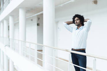 Young male entrepreneur with hands behind head looking away while contemplating in corridor at office - JOSEF02990