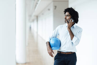 Junger männlicher Architekt mit Schutzhelm, der wegschaut, während er im Büro auf dem Flur mit seinem Handy telefoniert - JOSEF02992