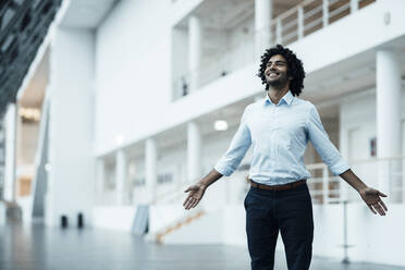 Smiling businessman standing with arms outstretched looking up in corridor at office - JOSEF03006