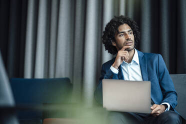 Thoughtful male entrepreneur looking away while sitting with laptop at office - JOSEF03020