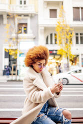 Woman with afro blond hair using smart phone while sitting on bench in city - OCMF01956