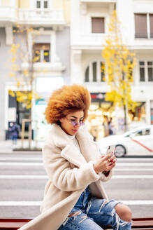 Frau mit afroblondem Haar benutzt ihr Smartphone, während sie auf einer Bank in der Stadt sitzt - OCMF01956