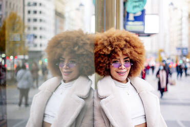 Smiling beautiful woman with afro hair leaning on glass wall in city - OCMF01966
