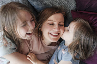 Glückliche Töchter, die ihre Mutter umarmen, während sie sich zu Hause auf die Couch legen - OGF00741