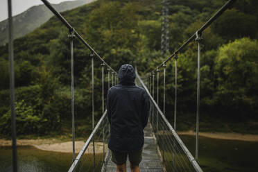 Woman wearing raincoat standing on suspension bridge over Sella river in forest - DMGF00420