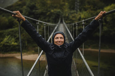 Lächelnde Frau in Regenmantel mit erhobenen Armen auf einer Hängebrücke über den Fluss Sella im Wald stehend - DMGF00421
