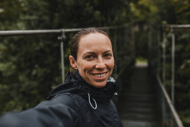 Lächelnde erwachsene Frau auf Hängebrücke an einem regnerischen Tag - DMGF00423